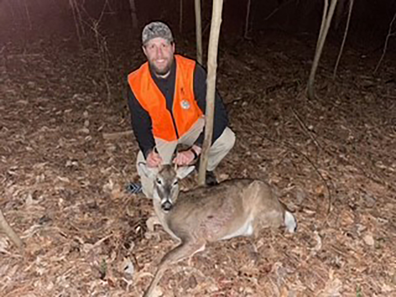 Una foto de un hombre con un chaleco naranja brillante arrodillado detrás de un pequeño ciervo de cola blanca en el suelo.