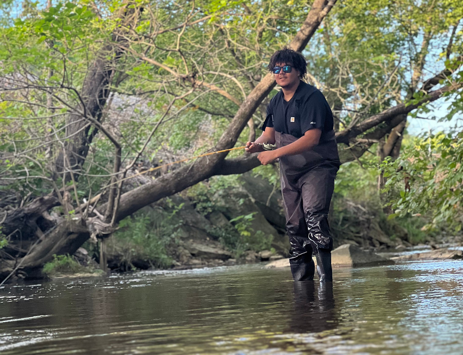 Una foto de un hombre en botas de agua parado hasta los tobillos en un arroyo con una caña de pescar.