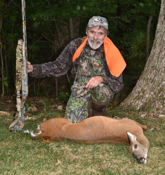Una foto de un hombre vestido de camuflaje y naranja sosteniendo una pistola y arrodillado detrás de una cierva de cola blanca en el suelo.