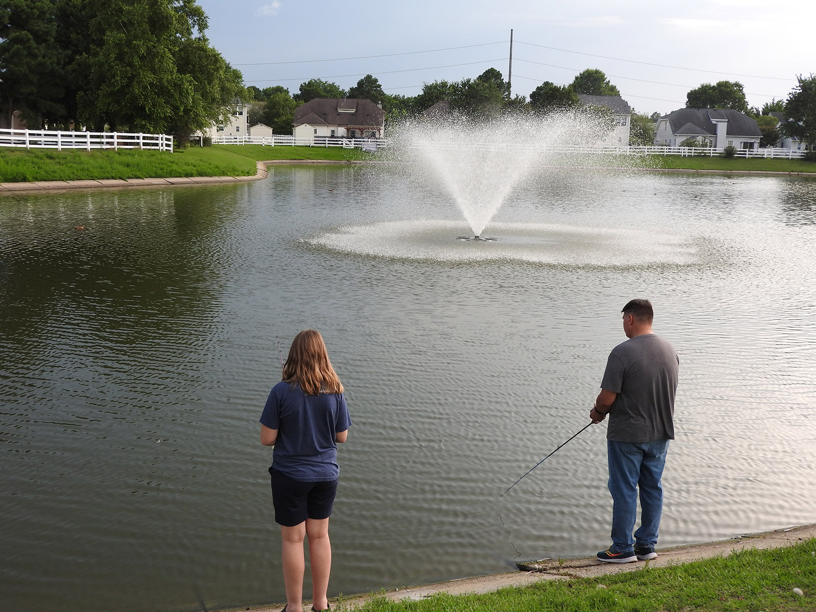 Una imagen de un hombre y una niña de pie en las orillas de un lago pescando en un estanque de retención local