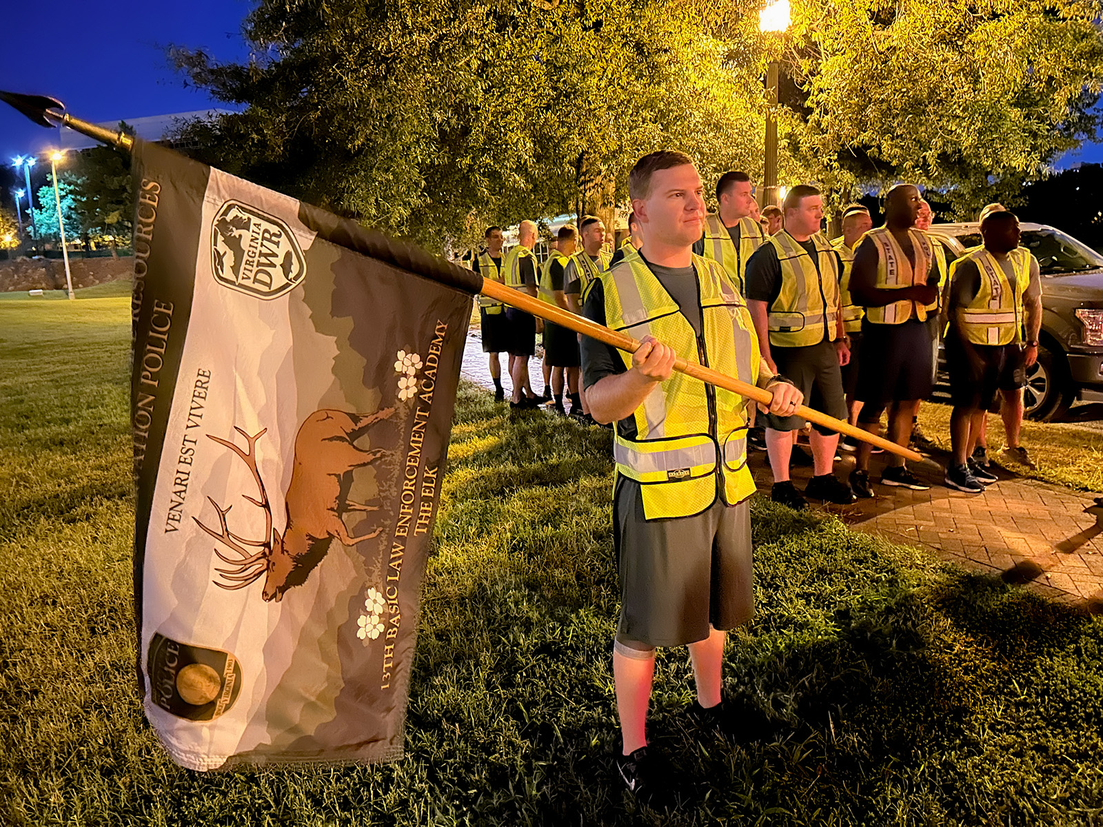 Una foto tomada a la luz del amanecer de un grupo de hombres y mujeres jóvenes preparándose para correr. El hombre de adelante sostiene una bandera de DWR con un alce en ella.
