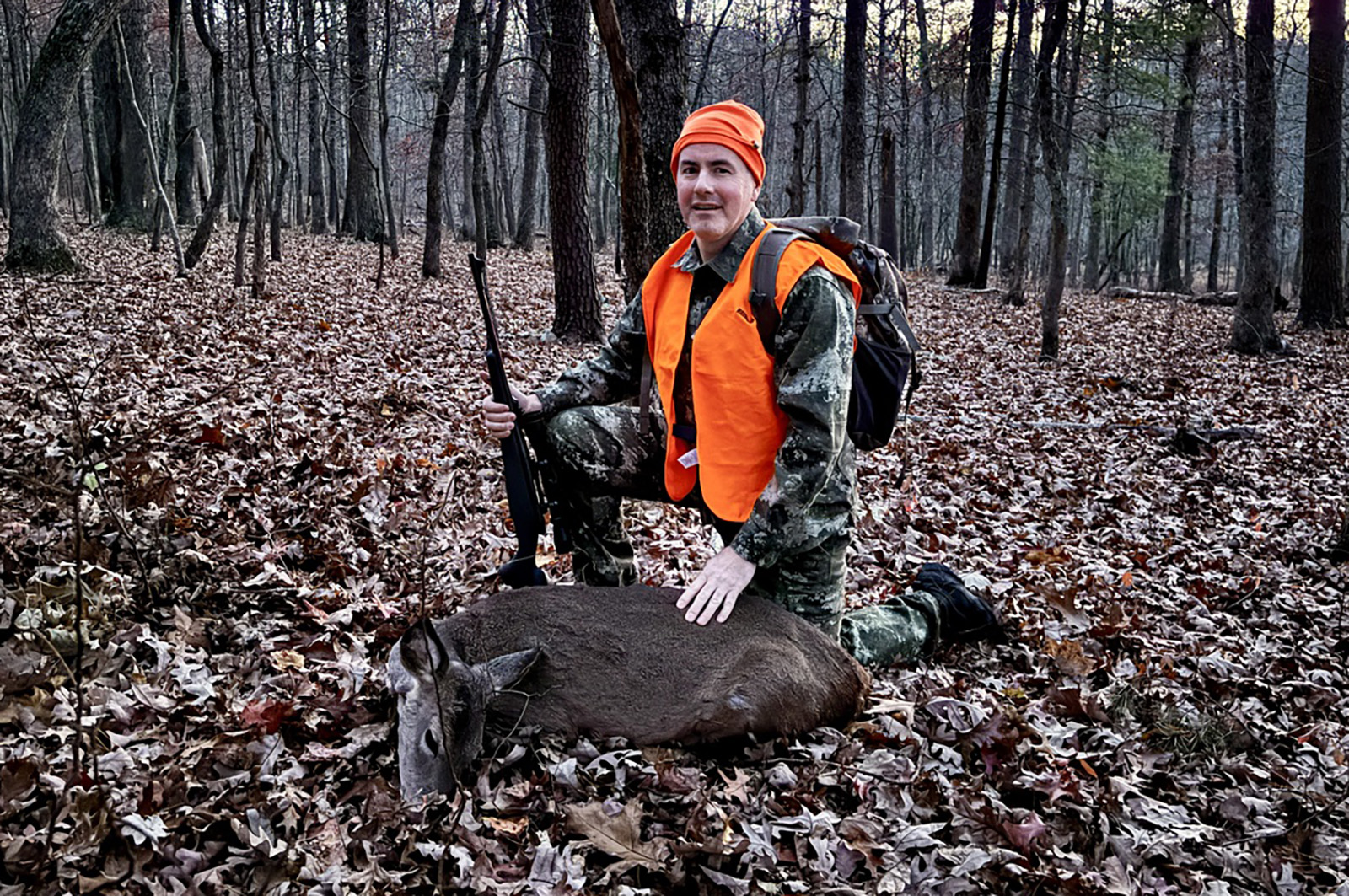 Una foto de un hombre con ropa de camuflaje y naranja de seguridad arrodillado detrás de una cierva cazada en el bosque.