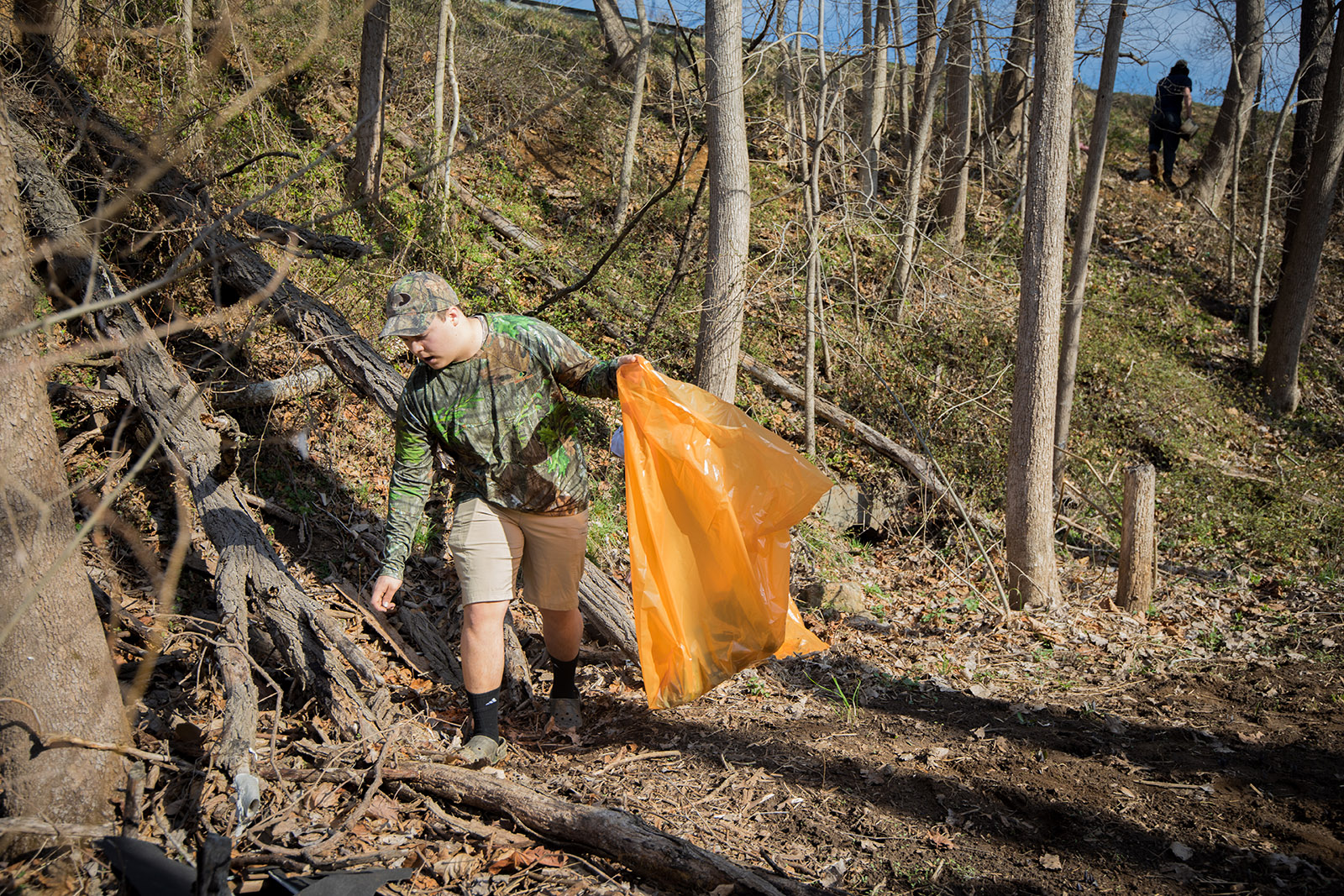Una imagen de un hombre camuflado con una bolsa de basura de color naranja brillante recogiendo basura en un bosque 