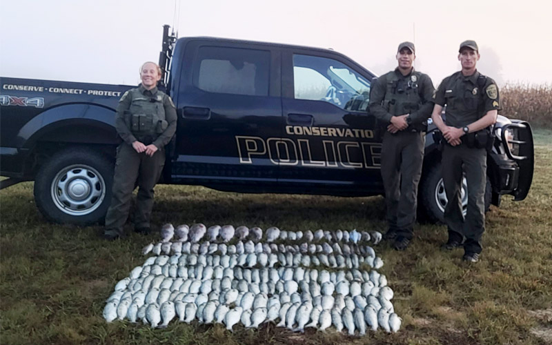 Una foto de tres oficiales uniformados de la Policía de Conservación parados frente a un camión CPO con una gran cantidad de peces en el suelo frente a ellos.