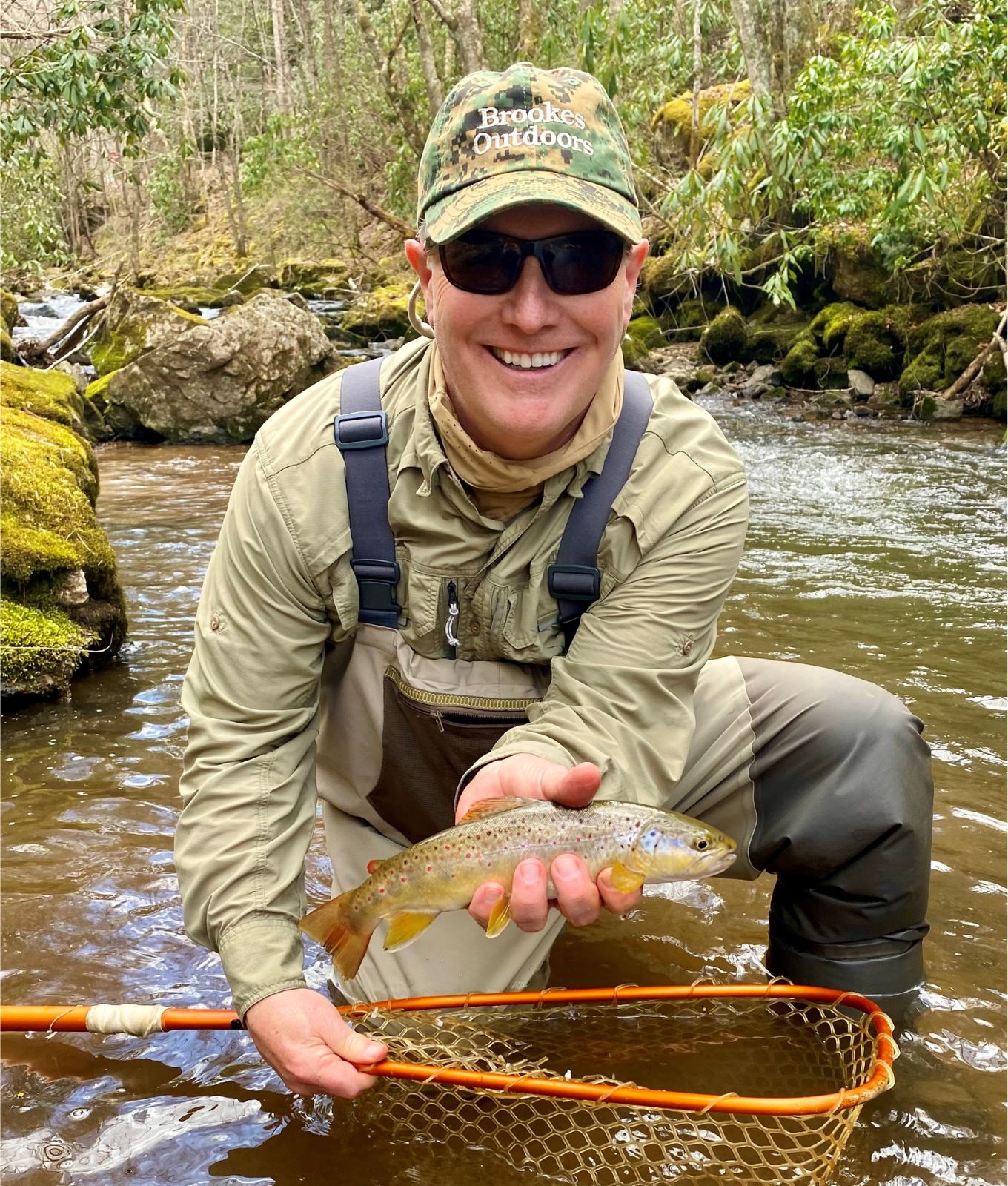 Una foto de un hombre sonriente en cuclillas en un hermoso arroyo, sosteniendo una red de pesca y una colorida trucha marrón. 