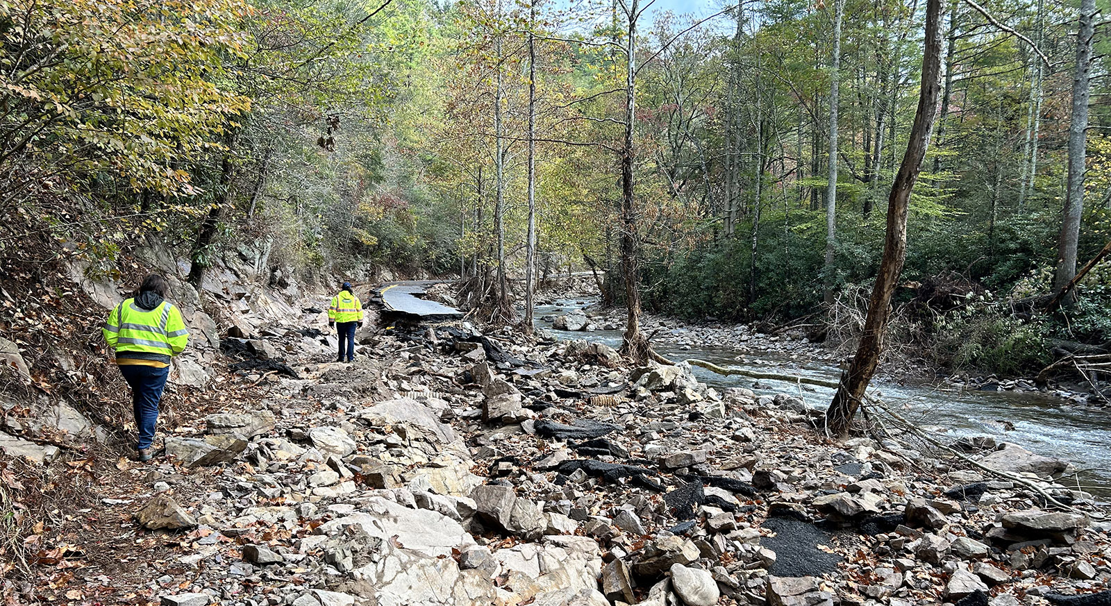 Una foto de dos personas con chalecos reflectantes caminando por una carretera destruida junto a un arroyo.