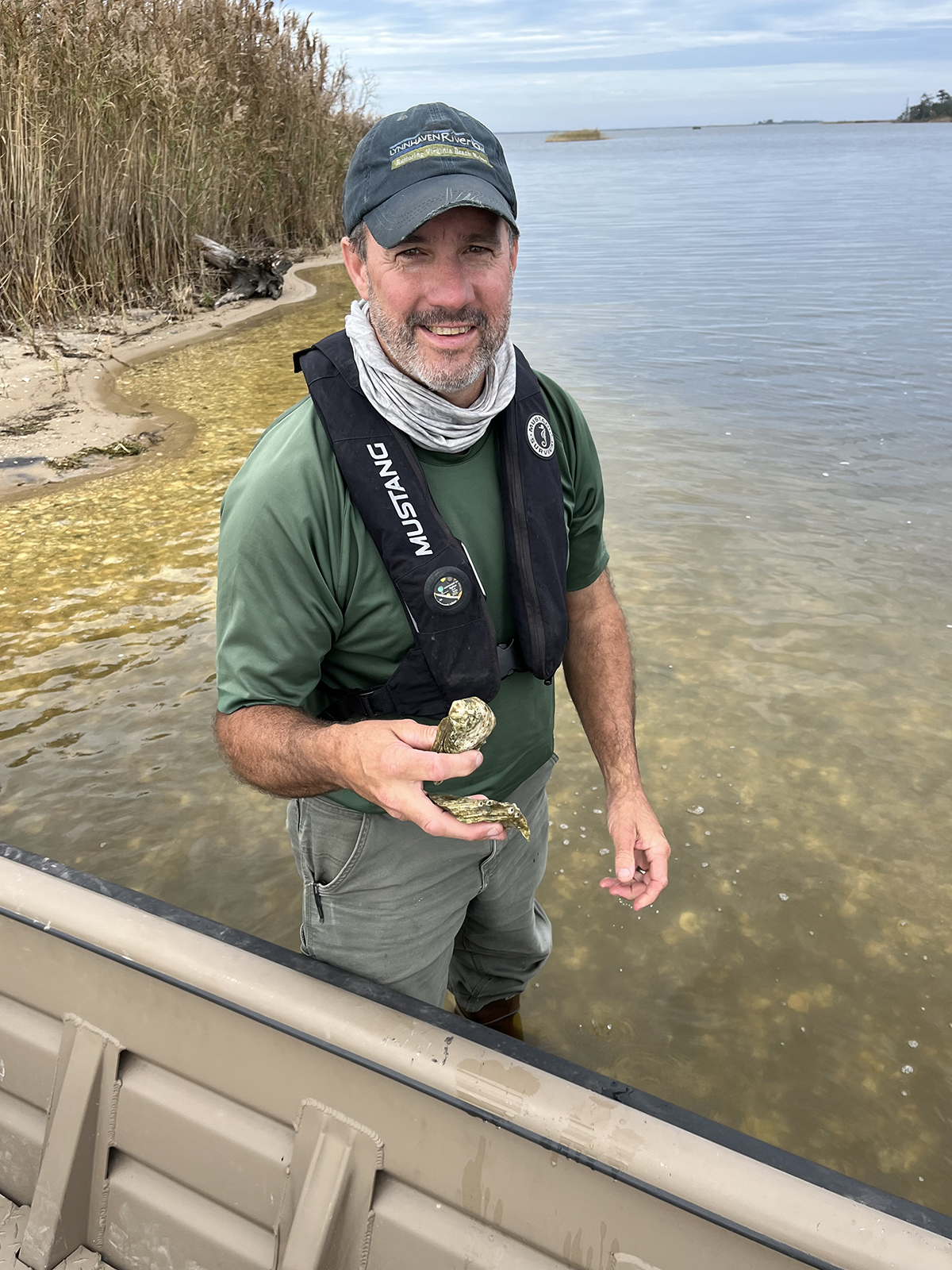Una foto de un hombre de pie con el agua hasta las rodillas junto a un bote, sosteniendo conchas de ostras.