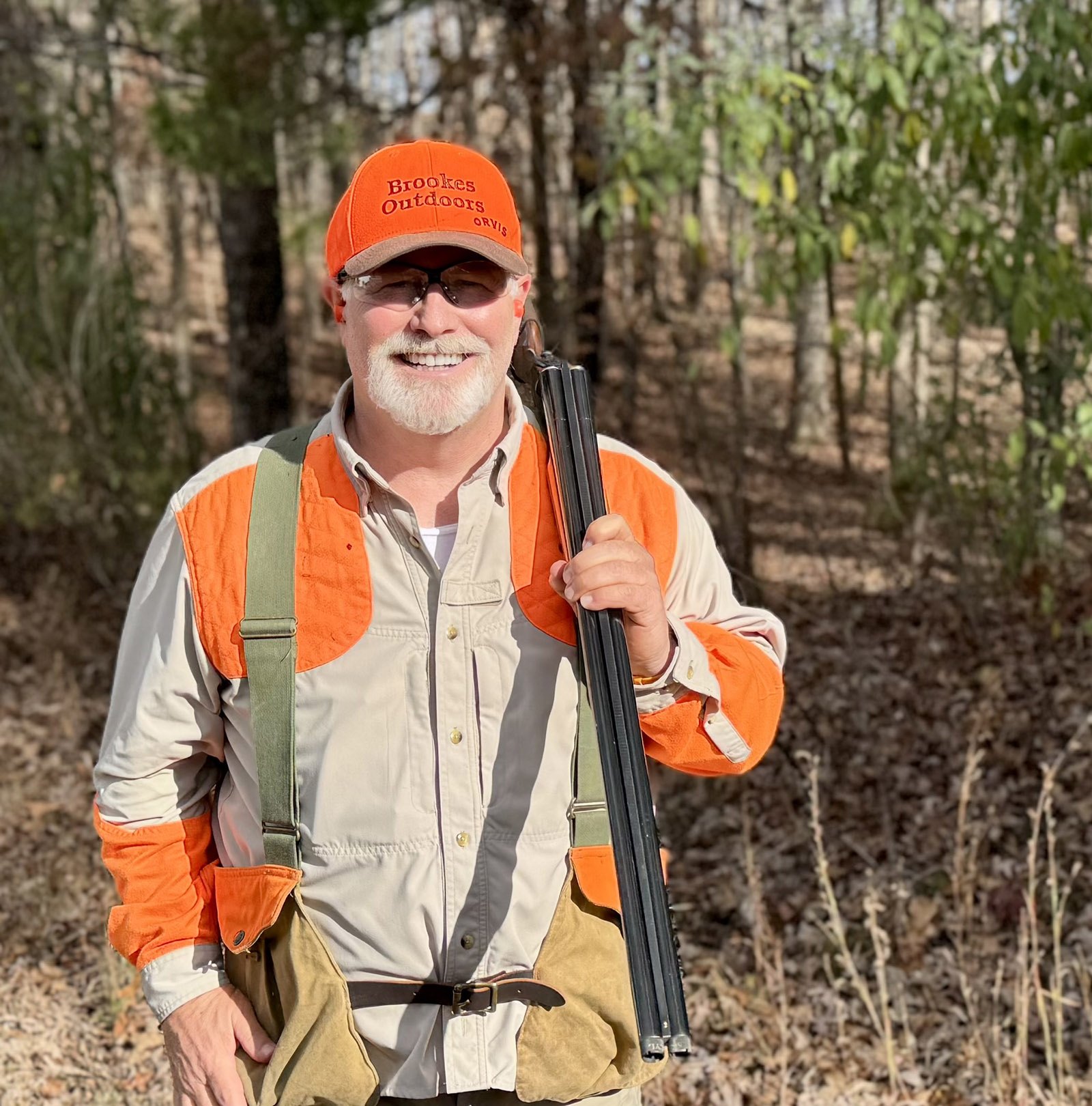 Una foto de un hombre con una escopeta al hombro, de pie en el bosque con un sombrero de color naranja brillante y una camisa con parches de color naranja fuego en los hombros y los brazos.