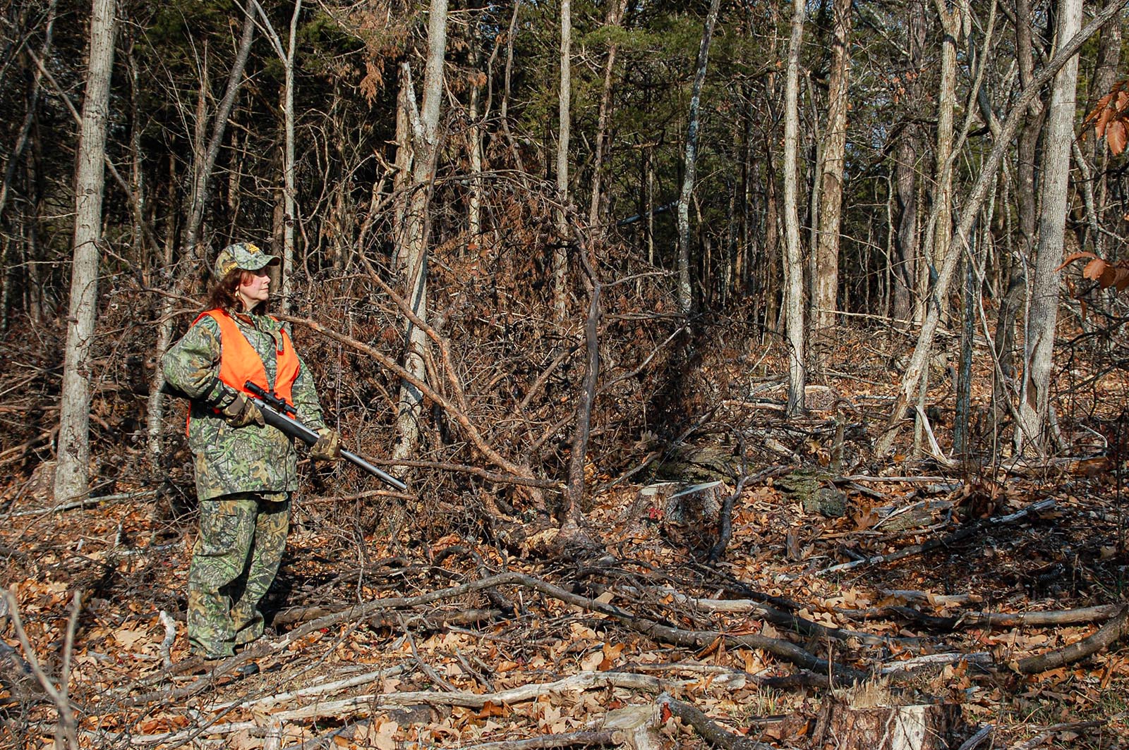 Una foto de un cazador sosteniendo una pistola, vestido de camuflaje y naranja brillante, de pie entre árboles cortados en un bosque.