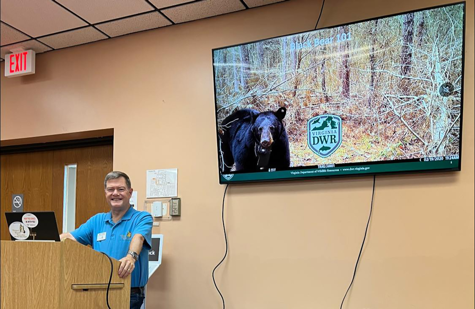 Una foto de un hombre detrás de un atril dando una presentación, con una foto de un oso negro en la pantalla detrás de él.