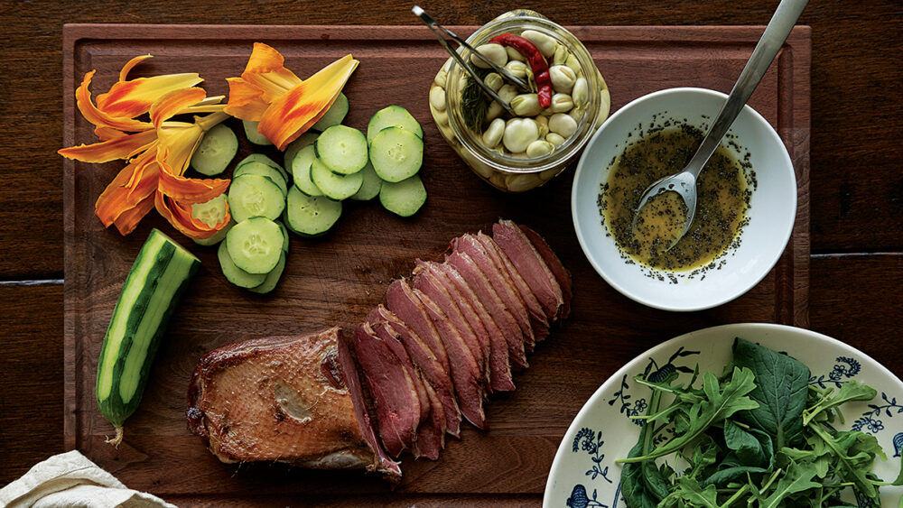 Una imagen de una porción de ganso ahumado, lirios de día, pepino, rúcula y habas en escabeche