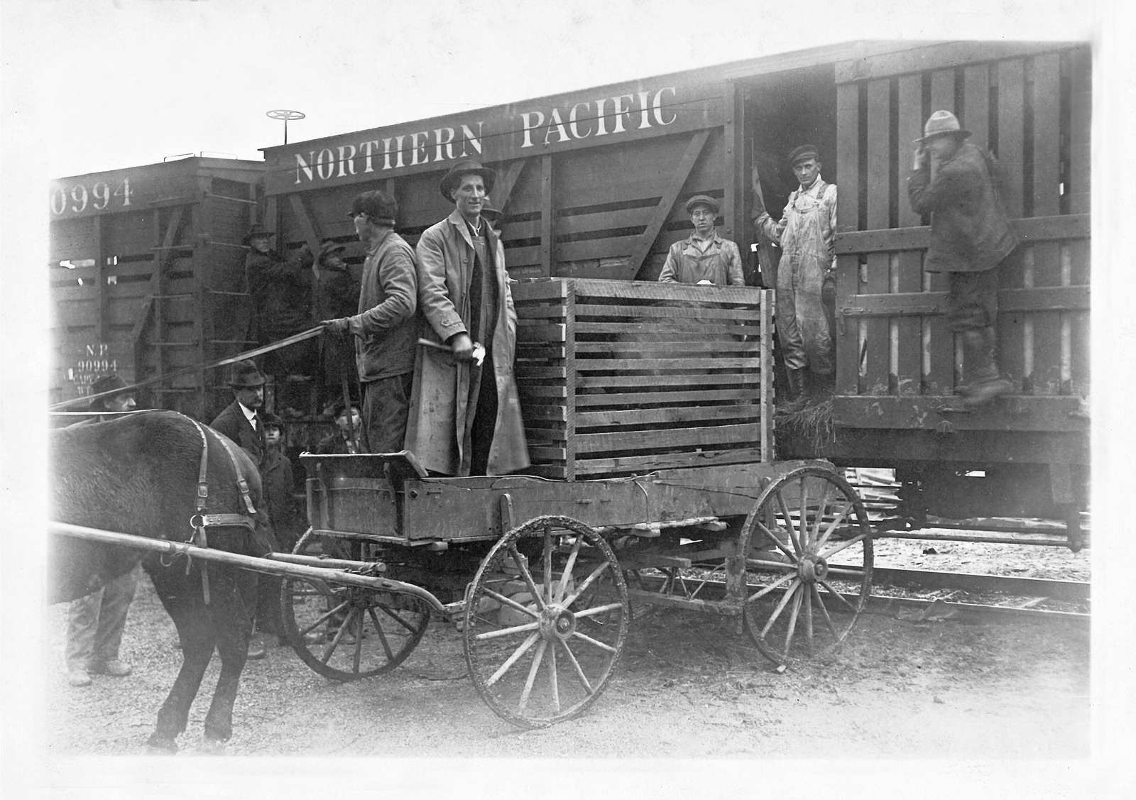 Una foto histórica en blanco y negro de cuatro hombres en un carro tirado por caballos con una gran caja respaldada por un vagón de ferrocarril.
