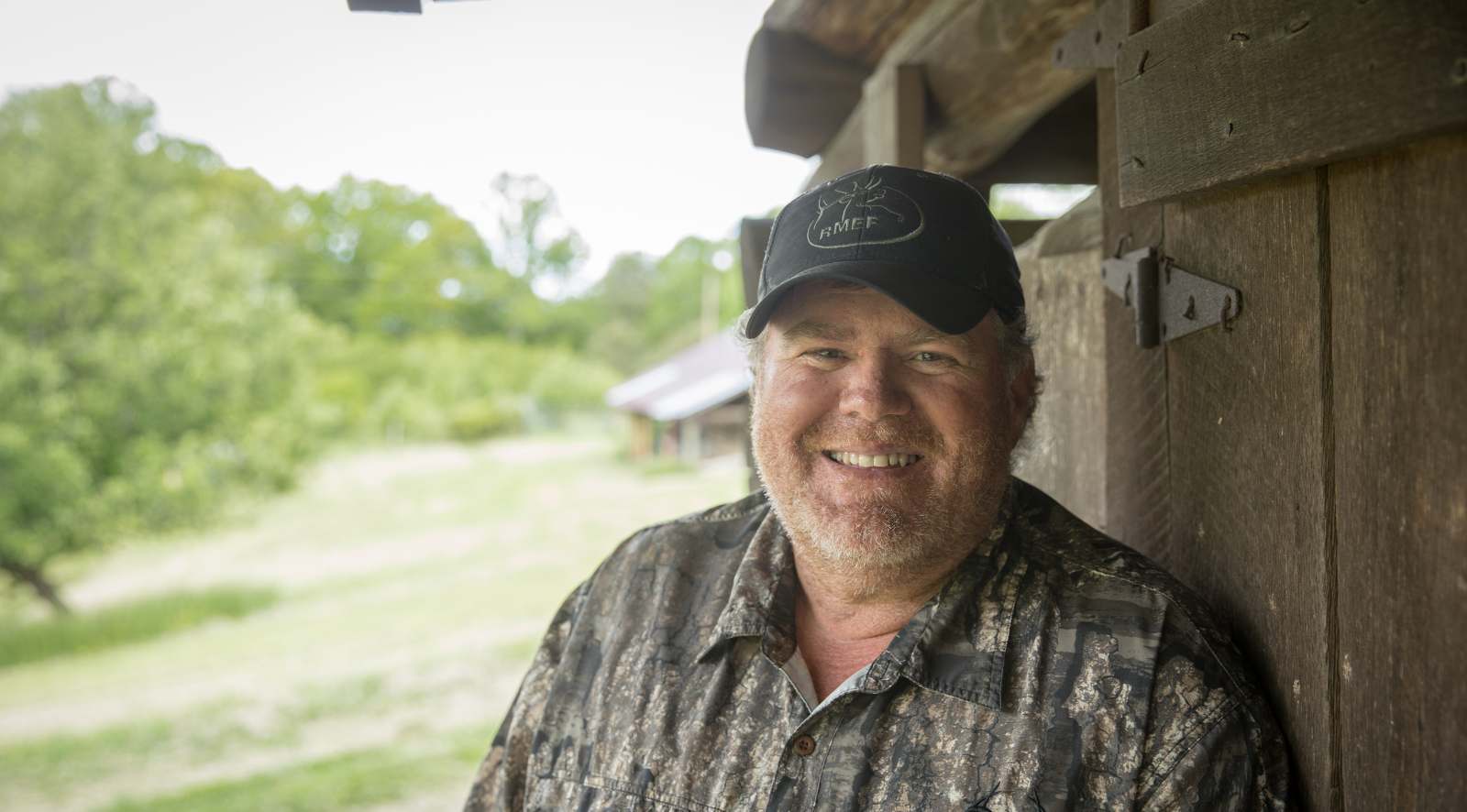 Una foto de retrato de un hombre con barba parado afuera junto a un edificio de madera tosca.