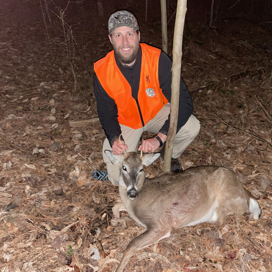 Una foto de un hombre vestido de naranja ardiente arrodillado con un ciervo de púas tirado en el suelo frente a él,