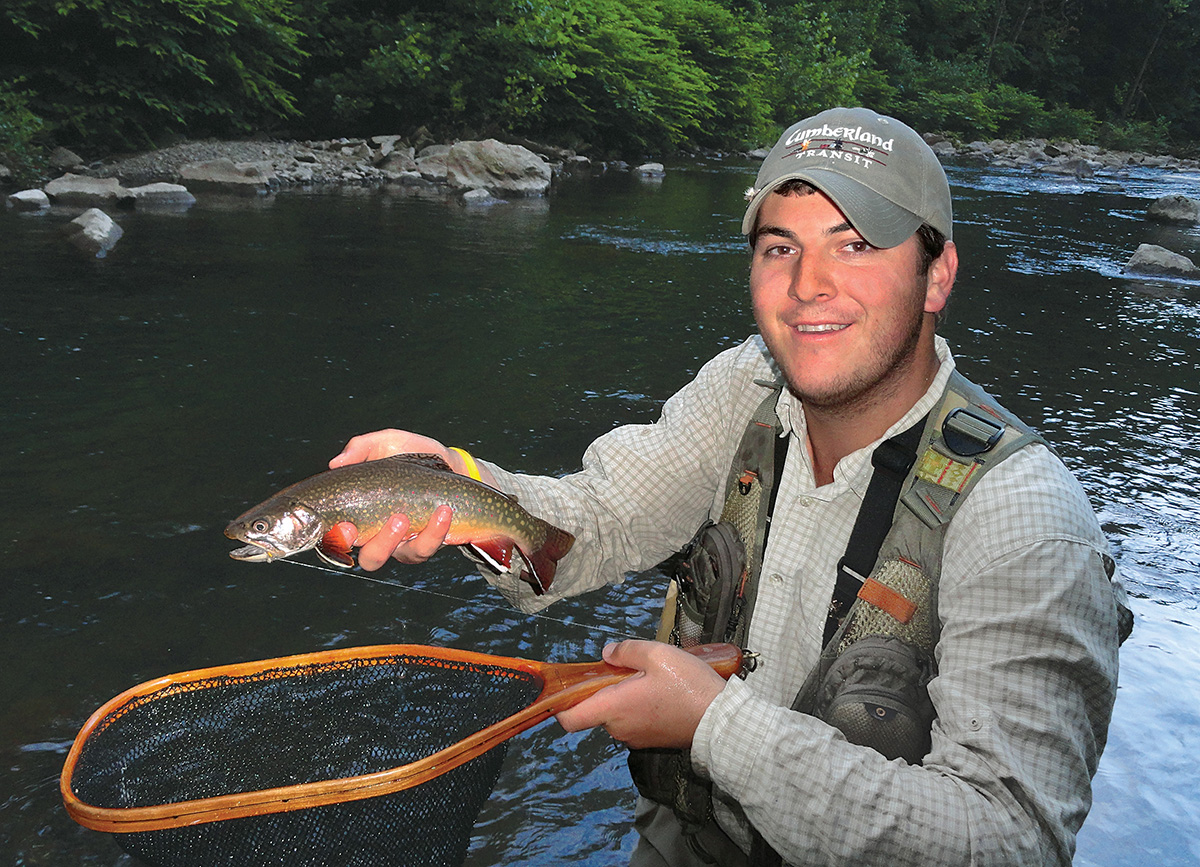 Una imagen de un pescador sosteniendo una red y una trucha marrón frente a un río