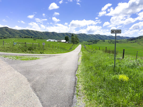 Un paisaje rural pintoresco con un camino pavimentado que se bifurca en dos direcciones. A la izquierda, la carretera conduce a un exuberante campo verde con colinas onduladas al fondo, mientras que a la derecha, la carretera continúa recto hacia una granja lejana. Un letrero que indica "608" es visible en el lado derecho de la carretera. El cielo es azul brillante con nubes blancas esponjosas esparcidas por todas partes.