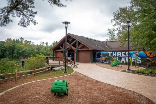 La entrada principal al Centro de la Naturaleza, que es un edificio marrón con un mural pintado en un lado que dice "Three Lakes." A la izquierda del edificio hay una plataforma de observación de madera con vista a uno de los lagos.