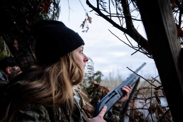 Una mujer vestida de camuflaje en una persiana de caza junto al agua