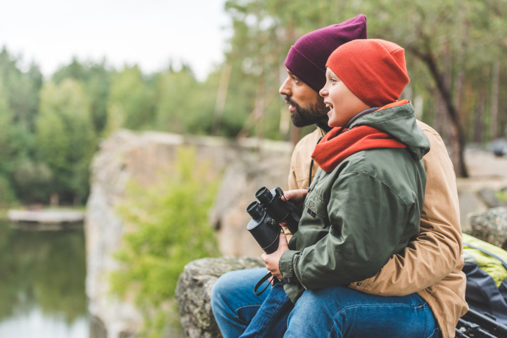 Una imagen de un hombre sosteniendo a un niño que sostiene binoculares, ambos individuos miran a la izquierda.