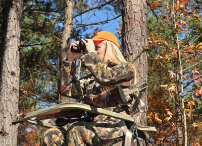 Una imagen de una mujer en una plataforma de observación de un árbol con un rifle y binoculares buscando cazar ciervos